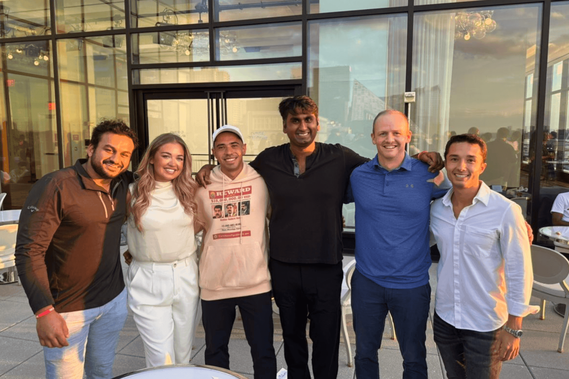 Group of six people standing together on a terrace, smiling during sunset.